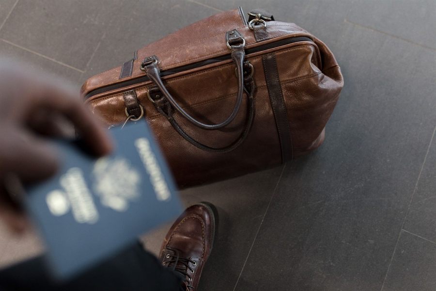 Hand holding a passport next to a brown leather travel bag